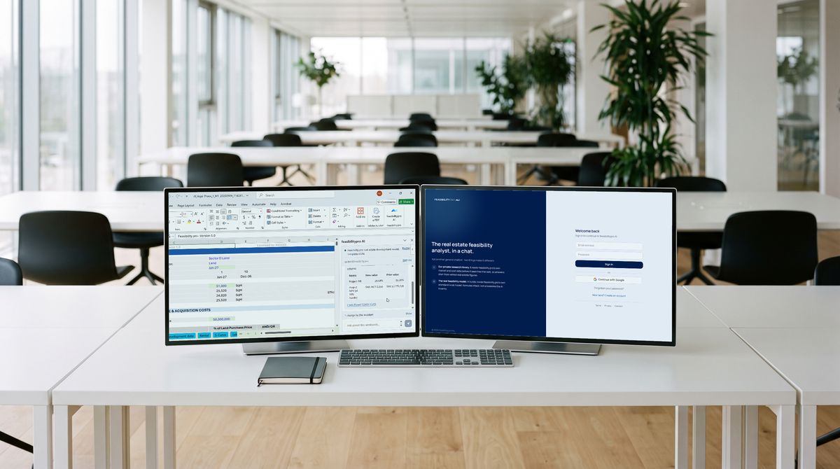 Two monitors on a desk in a large team workspace, with an assistant at work in a spreadsheet